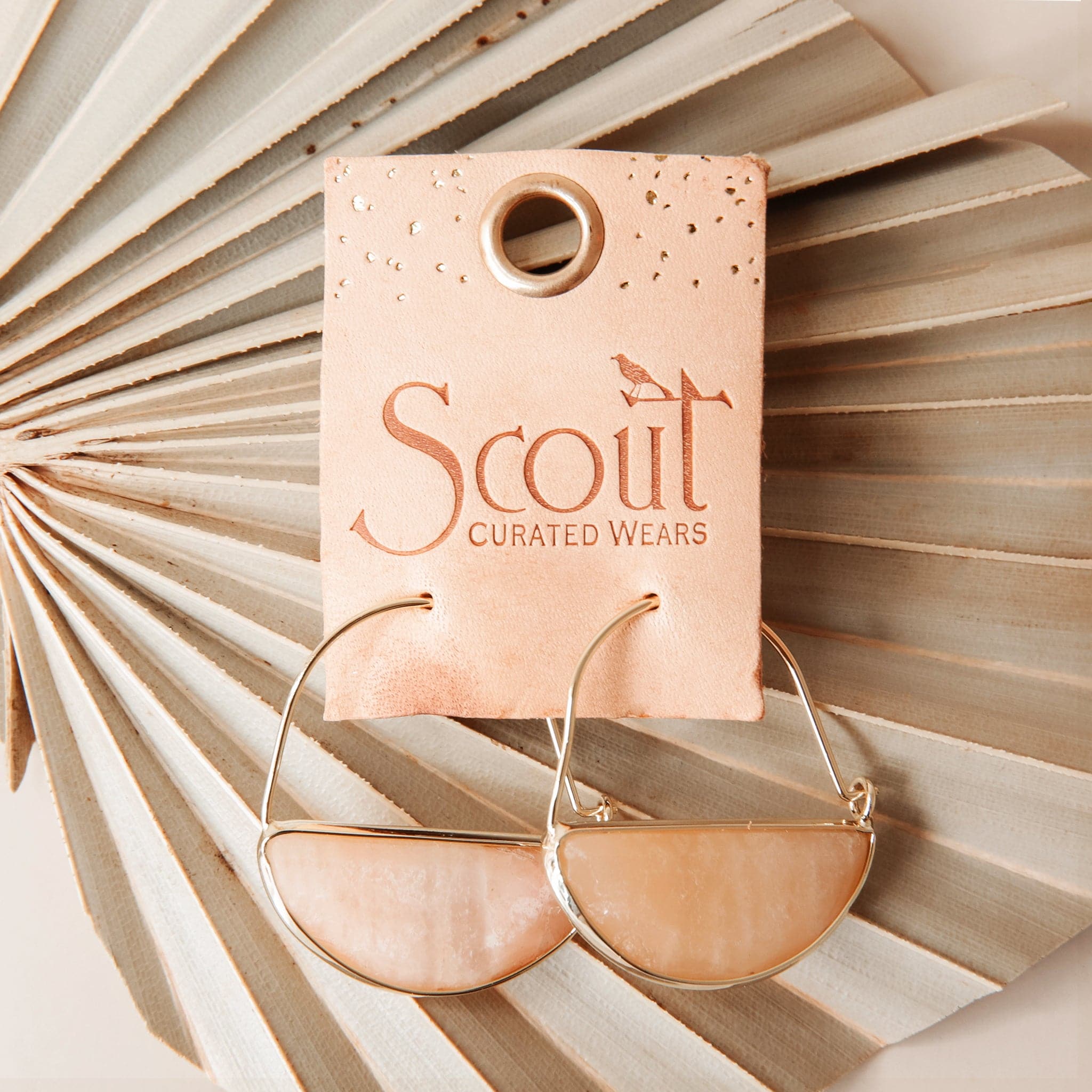 Laying on top of a dried palm frond is a pair of gold earrings. The top is a thin gold hoop. The bottom is a peach stone half circle with a gold border. The hoops are on a brown leather square holder. Etched in the middle the text reads ‘Scout curated wears.'