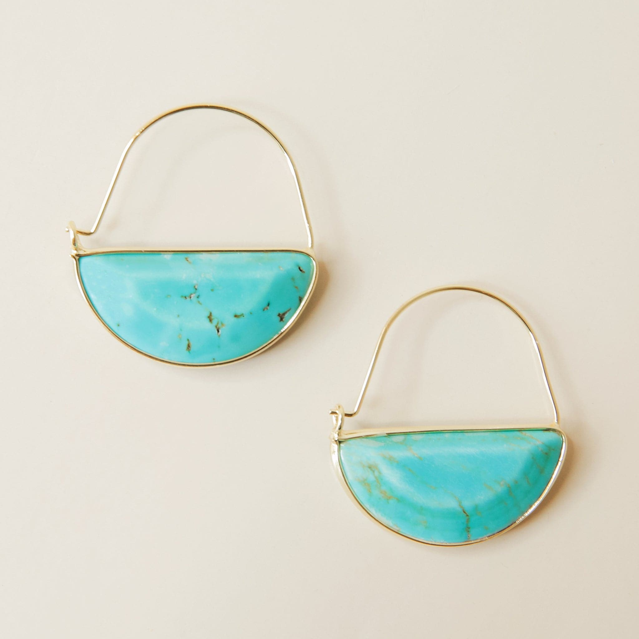 In front of a cream background is a pair of gold earrings. The top is a thin gold hoop. The bottom is a turquoise half circle with a gold border. 