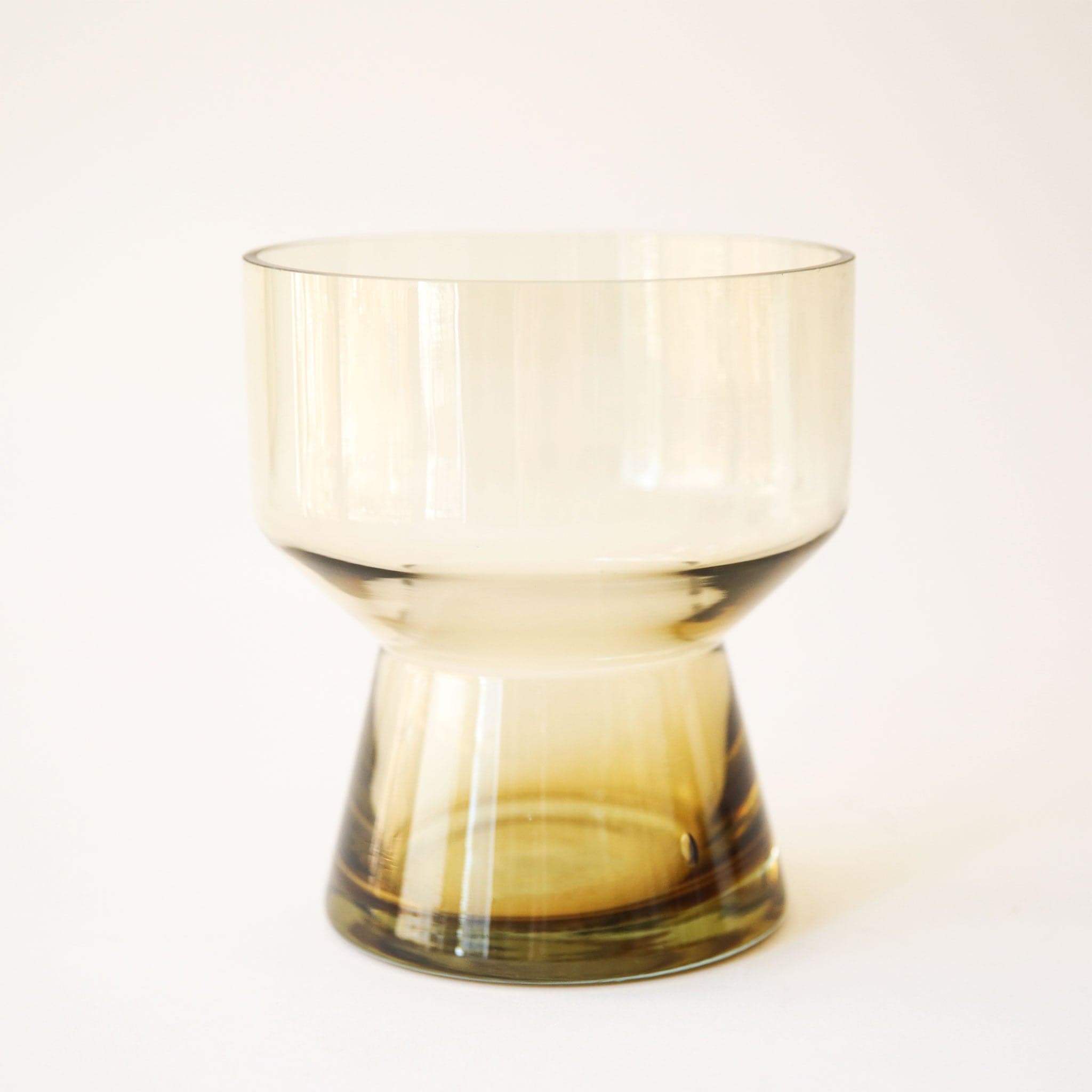 A compote style glass vase in an amber color in front of a white ground.