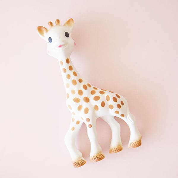In front of a soft pink background is a giraffe toy. The giraffe is white with tan spots and tan hooves. He has two black eyes, two brown ears and a two brown horns. 