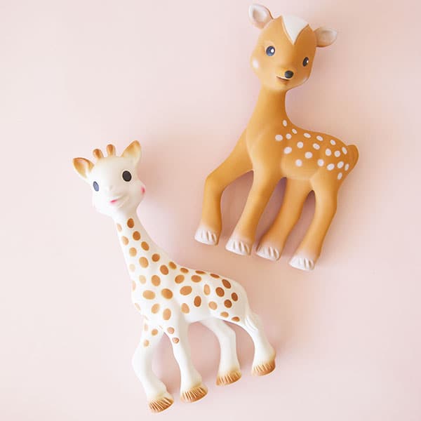 In front of a soft pink background is a giraffe and deer toy. On the bottom left corner is white giraffe with tan spots and tan hooves. He has two black eyes, two brown ears and a two brown horns. On the top right corner is a brown deer with white spots and white hooves. He has two black eyes and black nose. On top of his head he has two white ears and a white patch on his head. 