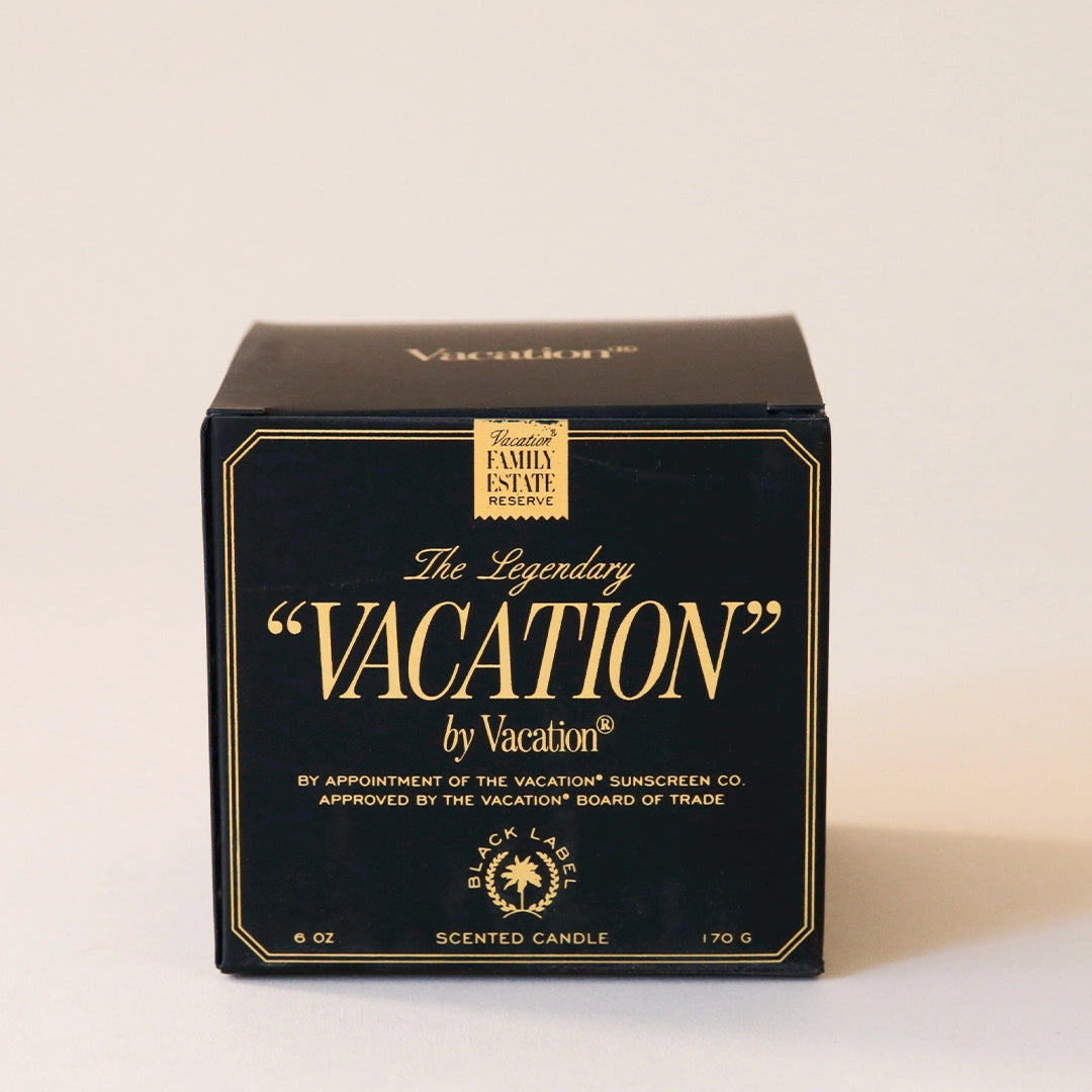 A black glass candle jar with a white soy wax blend on the inside. There is a gold label on the front of the candle that says, &quot;The Legendary &quot;Vacation&quot; by Vacation&quot;.