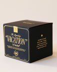A black glass candle jar with a white soy wax blend on the inside. There is a gold label on the front of the candle that says, "The Legendary "Vacation" by Vacation".