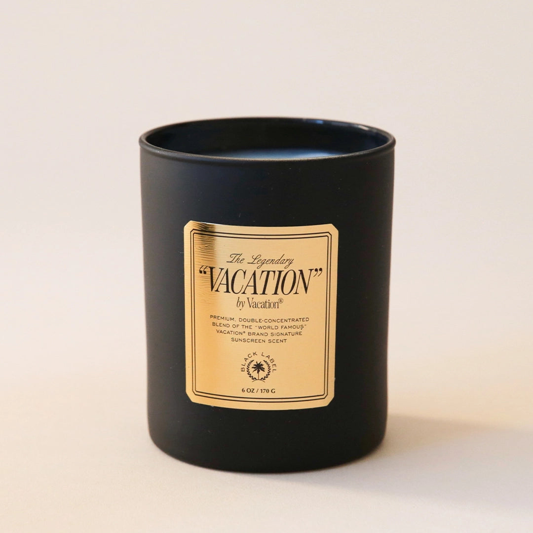 A black glass candle jar with a white soy wax blend on the inside. There is a gold label on the front of the candle that says, "The Legendary "Vacation" by Vacation".