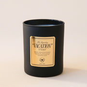 A black glass candle jar with a white soy wax blend on the inside. There is a gold label on the front of the candle that says, "The Legendary "Vacation" by Vacation".