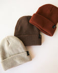 On a cream background is a rust colored ribbed beanie with a small black Brixton label on the edge of the folded brim photographed next to a cream and dark brown beanie.
