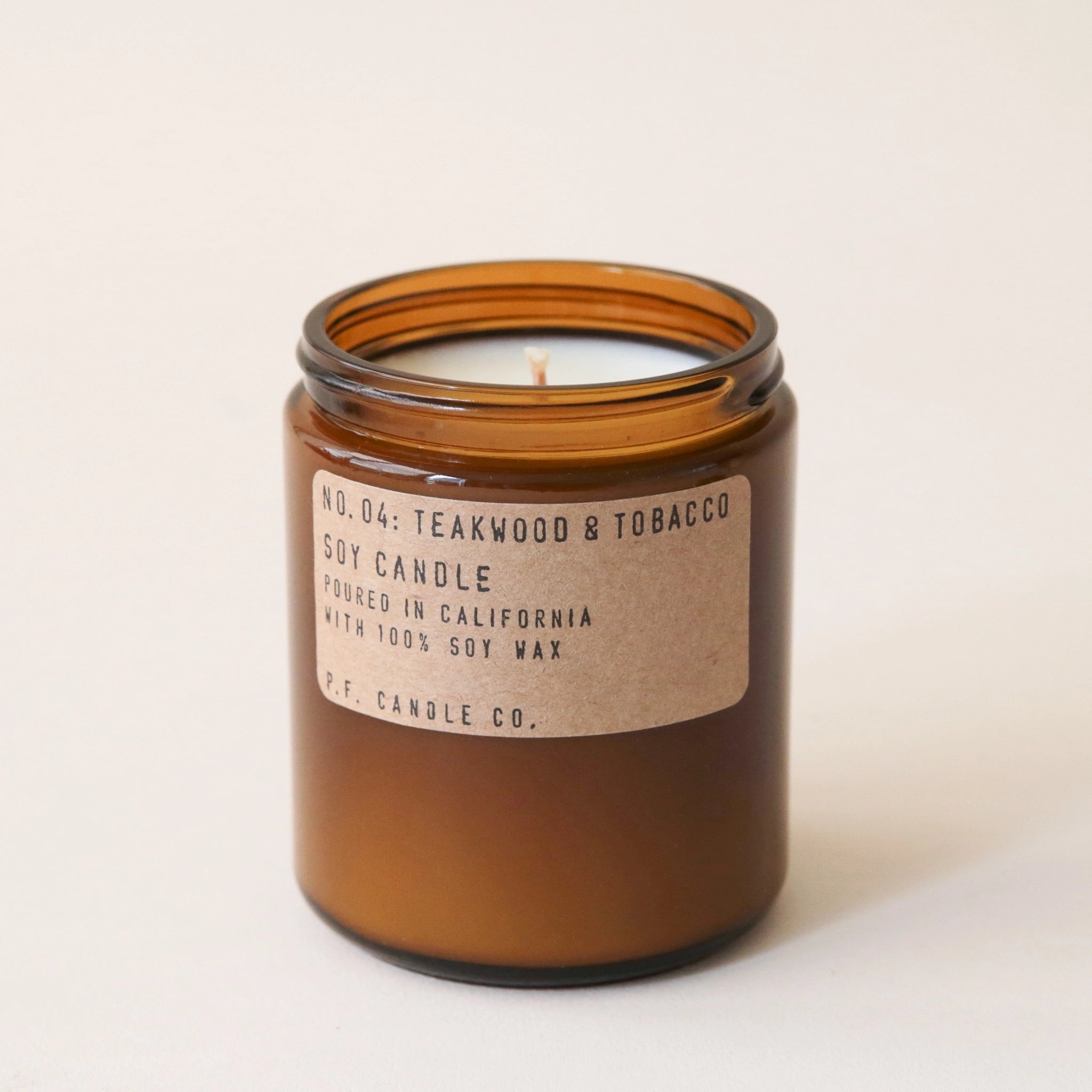 Against a white background is a dark amber, round jar. On the front of the jar is a brown, rounded, rectangular sticker with black text. The text reads ‘no.04 teakwood &amp; tobacco. Soy candle. Handmade in California with 100% soy way. P.F. candle co.’