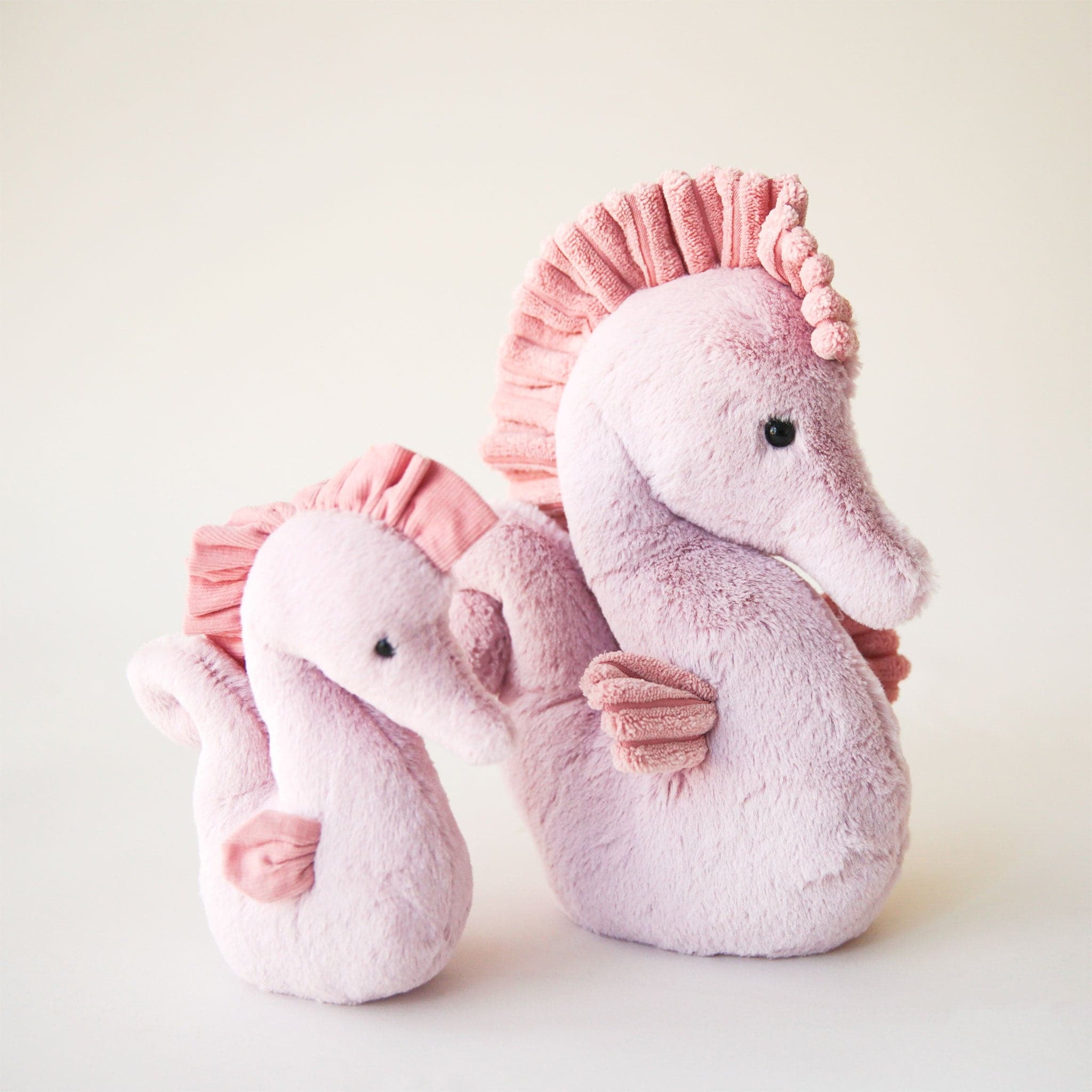 On a cream background is two different sized pink seahorse stuffed animals with a sweet face. 