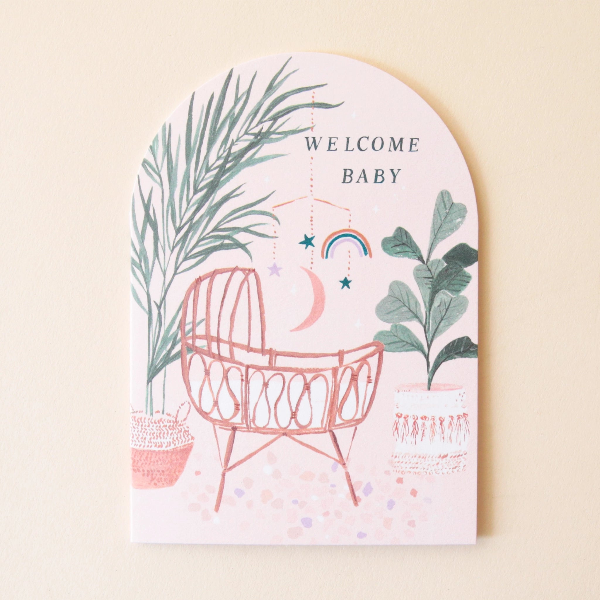 An arched greeting card with a sunset desert color pallet along with an illustration of a woven rattan baby&#39;s crib along with bohemian illustrations of lush natural foliage, bamboo, palm trees, and dried flowers and text at the top that reads, &quot;Welcome Baby&quot; in black letters.