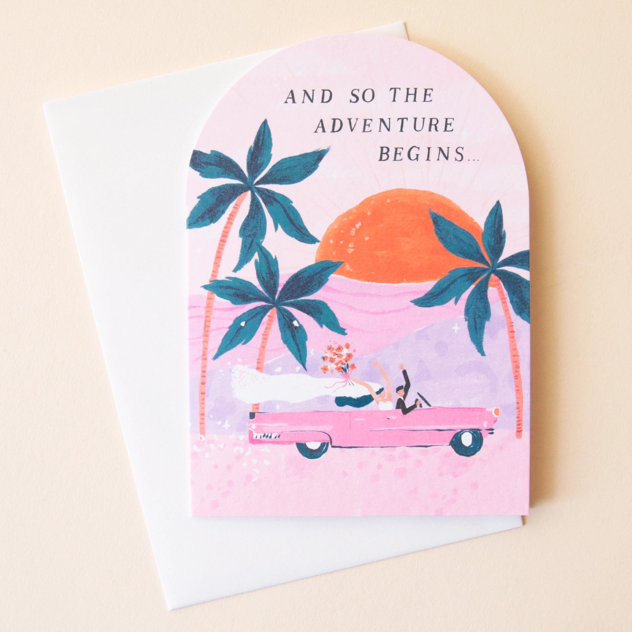 A pink arched greeting card featuring three palm tree illustrations along with a pink vintage car riding into the sunset with a bride and groom holding their hands and bouquets in the air as well as text that reads, "And So The Adventure Begins..." in black letters.