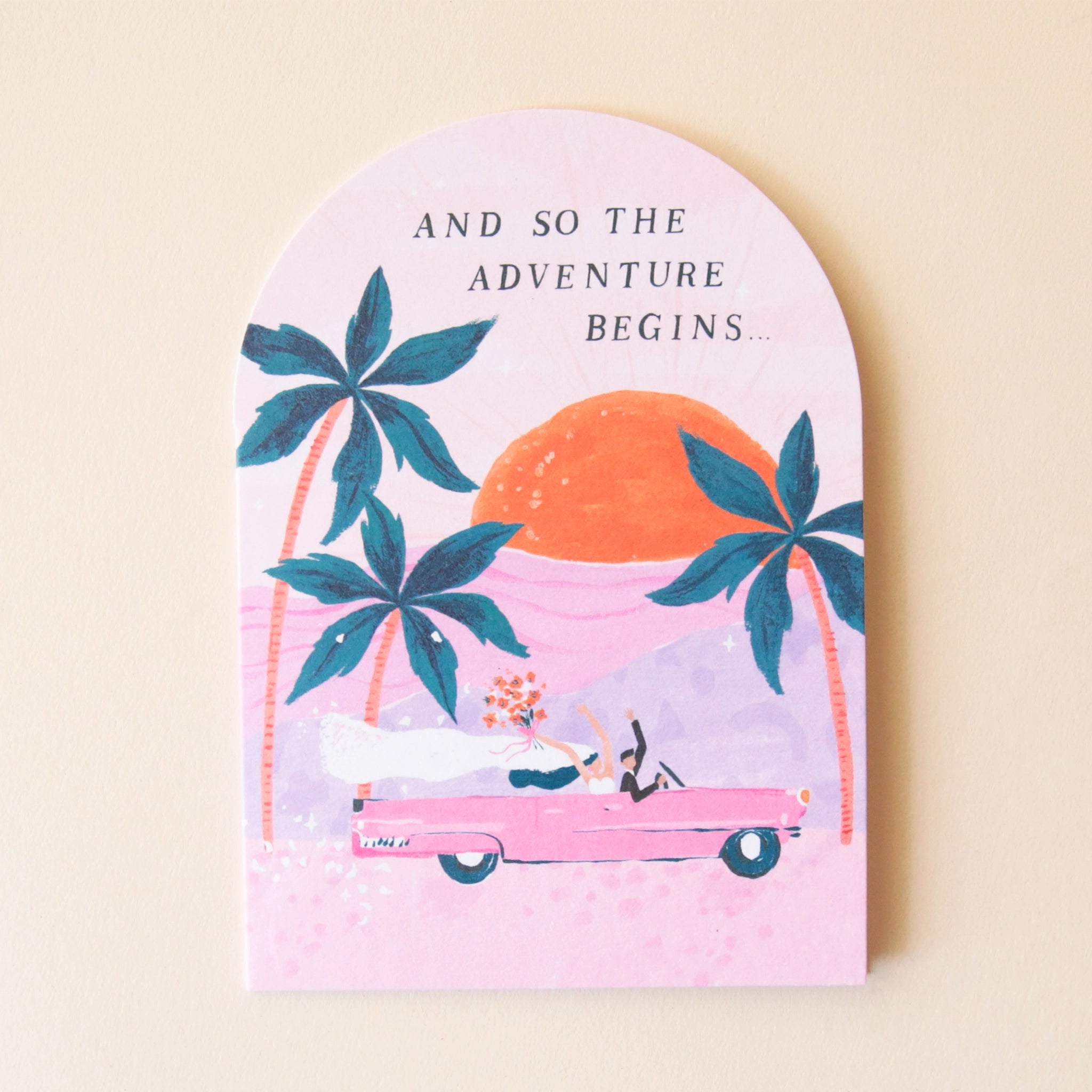 A pink arched greeting card featuring three palm tree illustrations along with a pink vintage car riding into the sunset with a bride and groom holding their hands and bouquets in the air as well as text that reads, &quot;And So The Adventure Begins...&quot; in black letters.
