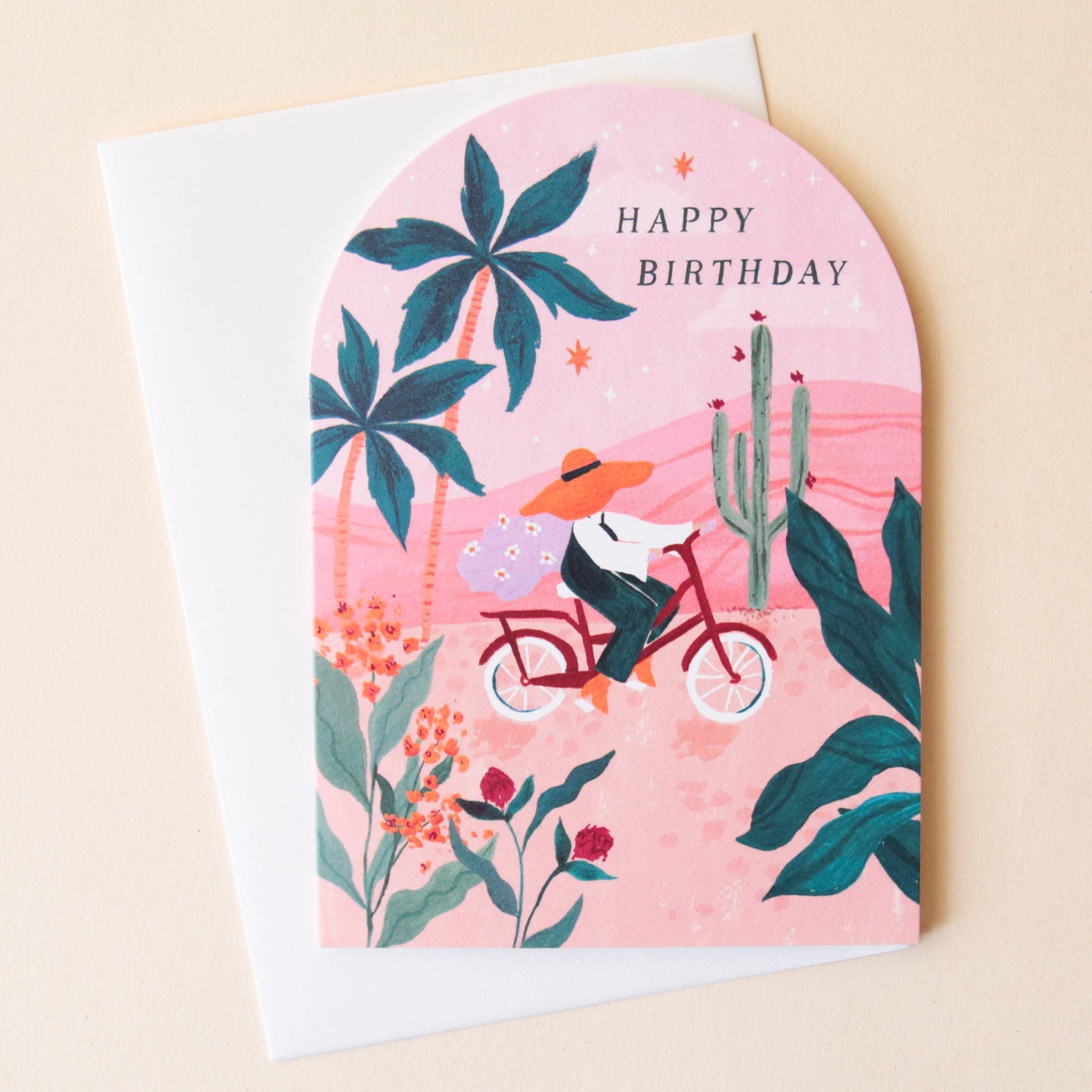 A pink arched greeting card with beautiful illustrations of palm trees, cacti and florals along with someone riding their red bicycle through the foliage along with text that reads, "Happy Birthday" in black letters.