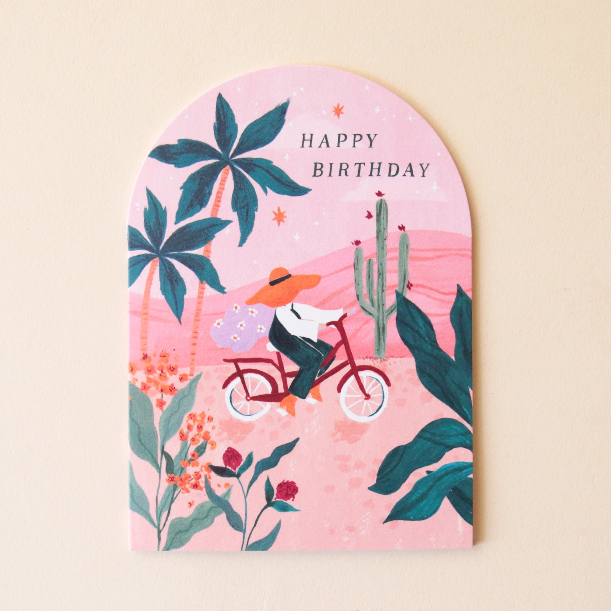 A pink arched greeting card with beautiful illustrations of palm trees, cacti and florals along with someone riding their red bicycle through the foliage along with text that reads, "Happy Birthday".