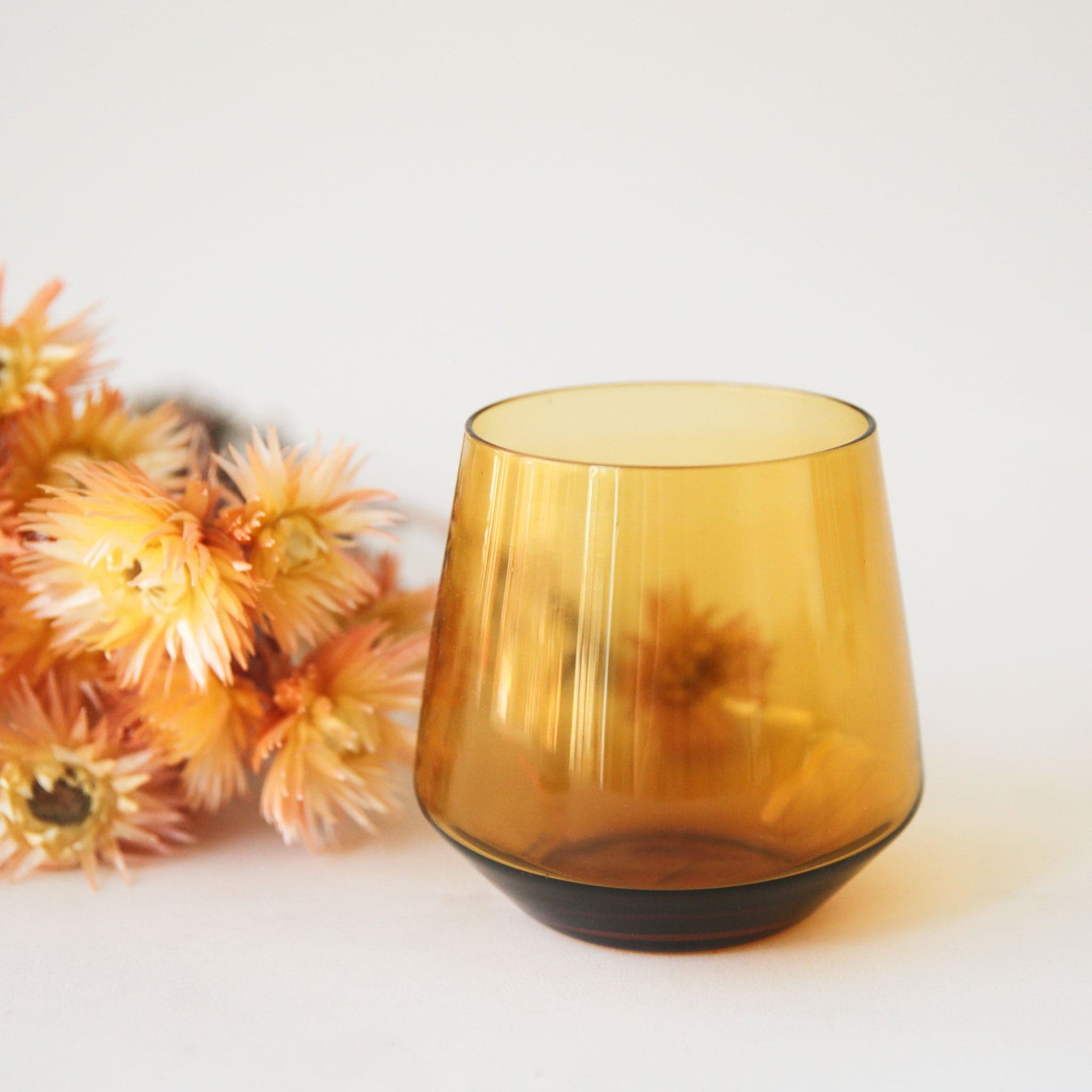 An amber colored stemless drinking glass with an angled / slightly curved bottom edge and staged next to orange dried florals.
