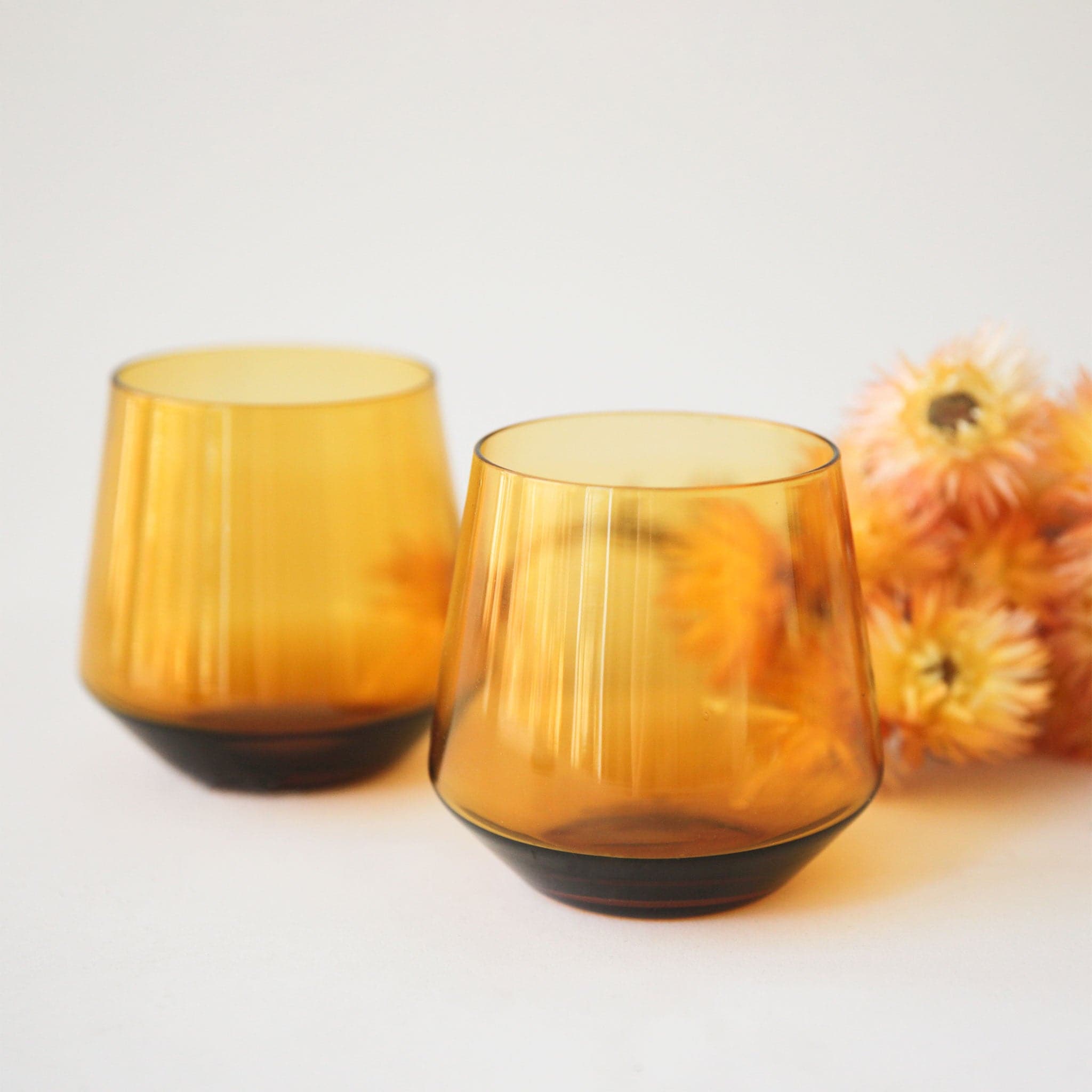 Amber colored stemless drinking glasses with an angled / slightly curved bottom edge and staged next to orange dried florals.