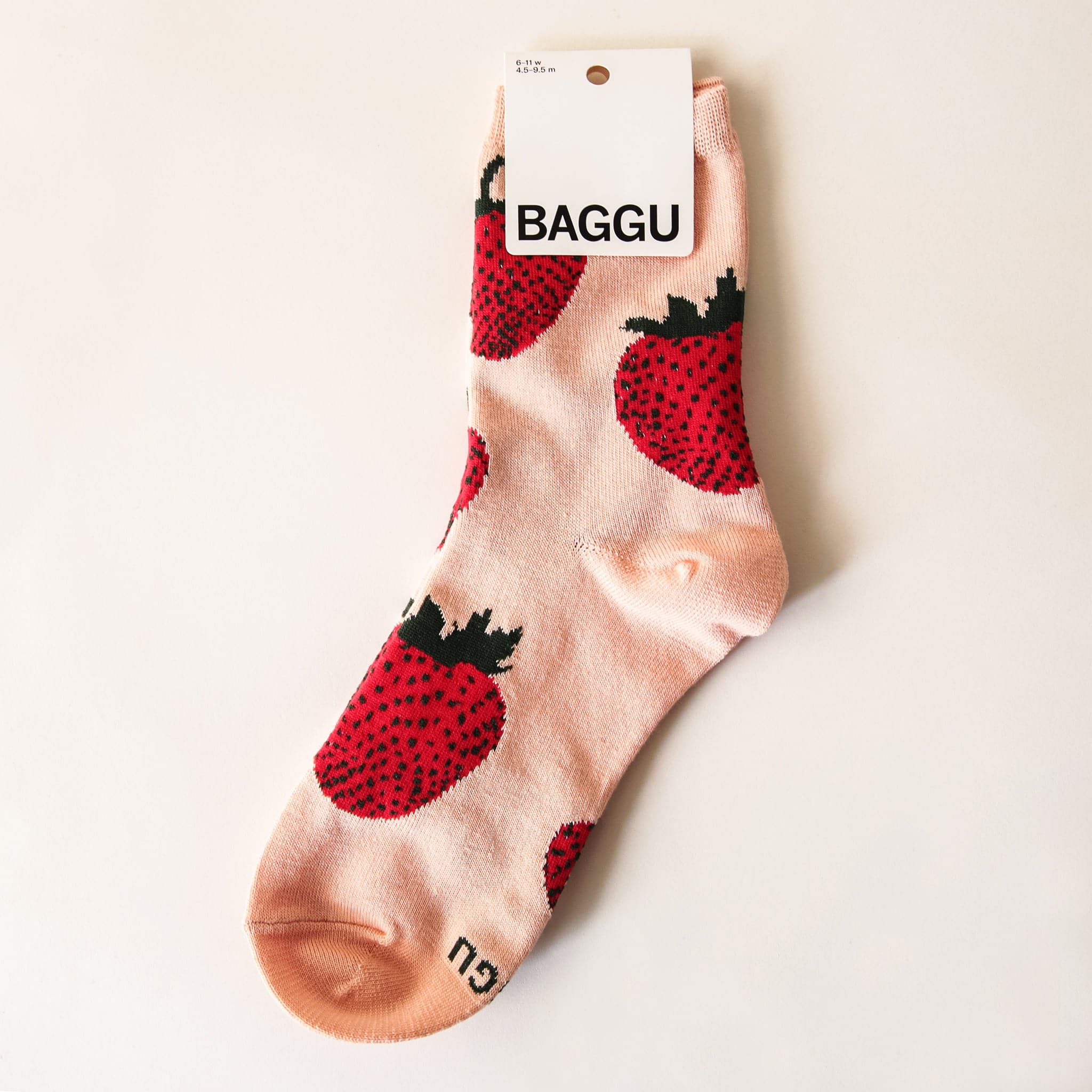 In front of a tan background is a pink, ankle sock with red strawberries on it. The strawberries have black dots and green leaves on top. At the top of the sock is a white tag with black text that reads ‘Baggu.’ 
