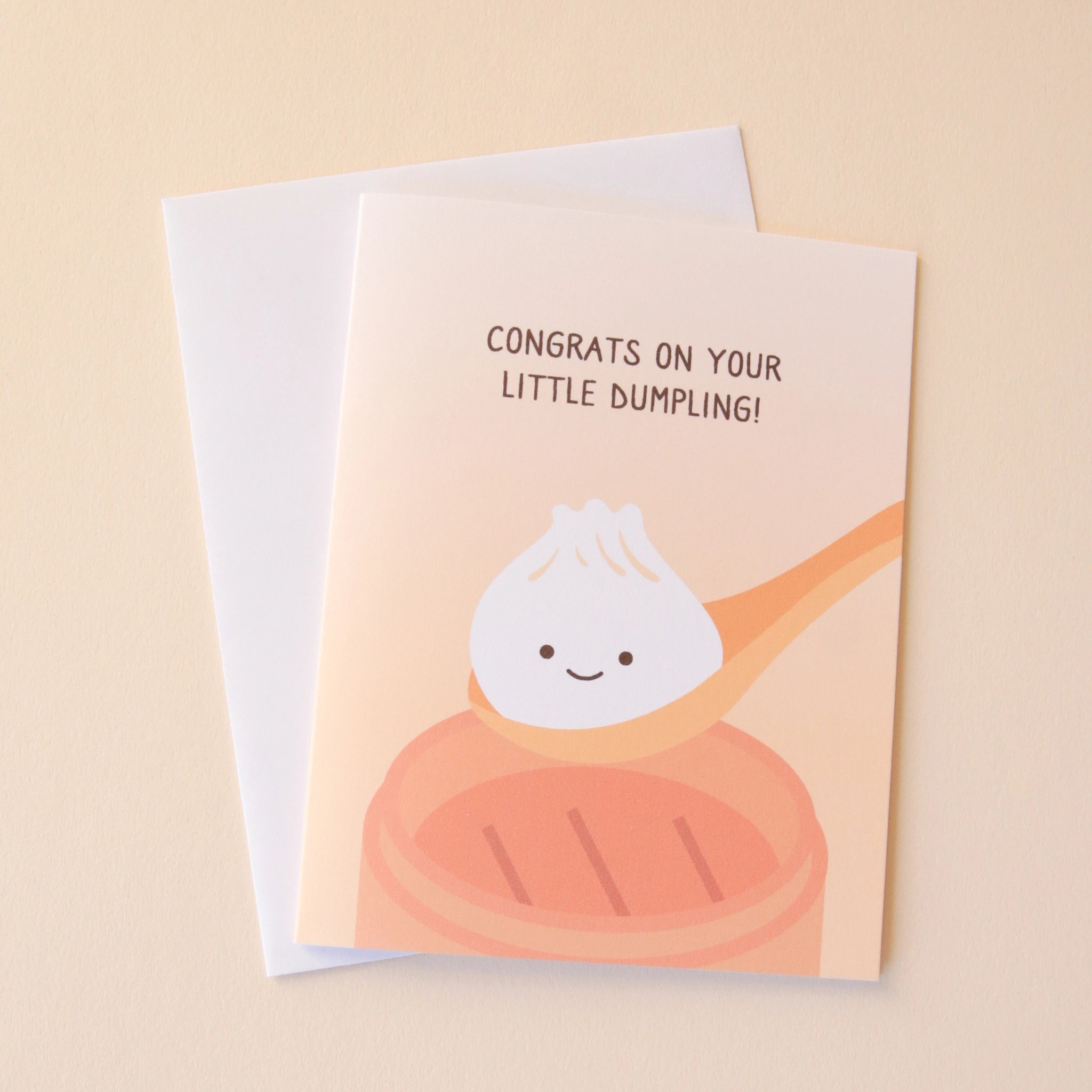 A light orange card with a illustration of a dumpling in a spoon with a smiling face along with text above it that reads, &quot;Congrats On Your Little Dumpling!&quot; in dark brown letters along with a white envelope.