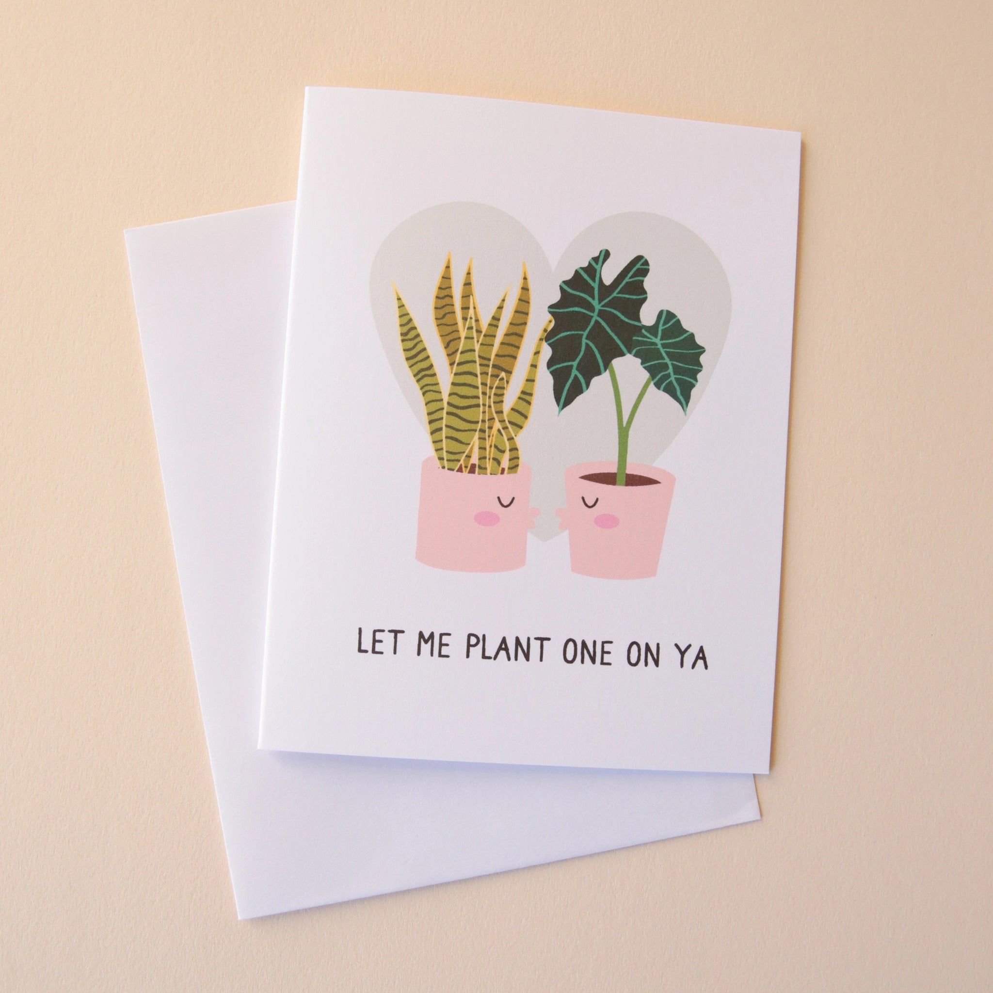 A white card with a light grey heart in the background behind a snake plant and an Alocasia Polly that are planted in two separate pink pots that have kissing faces and are going in for a kiss. There are black words underneath that read, "Let Me Plant One On Ya".