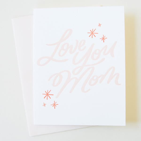 A white card with &quot;Love You Mom&quot; in light pink cursive text. It also has three red star details above and below the text.