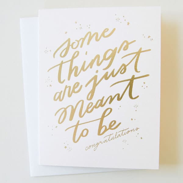 A cream card with gold foiled words that read, &quot;Some things are just meant to be. Congratulations&quot; alongside a matching white envelope. 