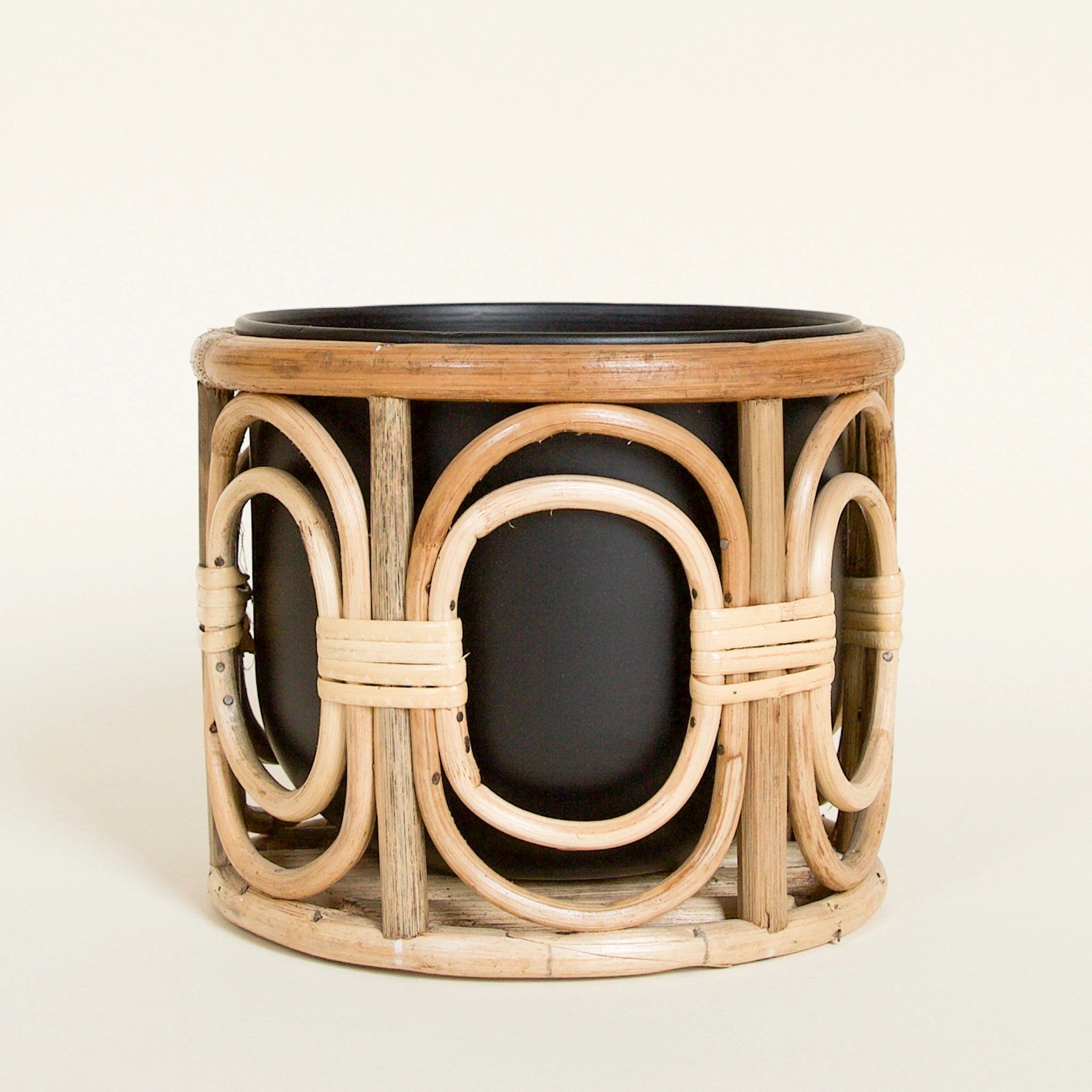 A table top rattan plant stand with black metal insert pot on a white background. Stand features bent wood layered open ovals that wrap around the pot and are separated by vertical lines of wood.