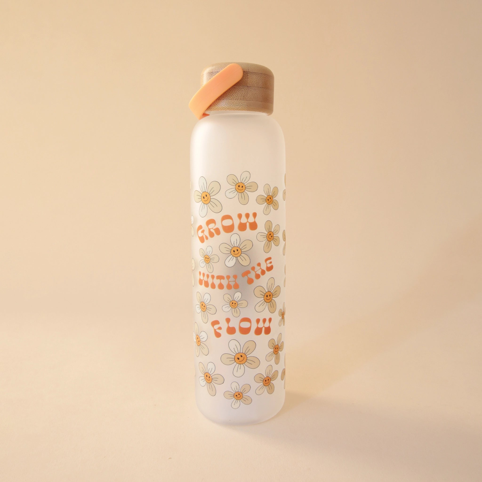 A tall and thin glass water bottle with a wood lid a tangerine handle and text on the front that reads, "Grow With The Flow" in groovy orange letters as well as smiley face daisy graphics all around the the words.
