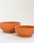Two terracotta orange pedestal bowl planting pots, both vessels are round and have tapered bases. The one in front is the smaller of the two and the pair sit empty besides each other.