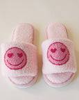 Pink fuzzy slide slippers with a darker pink heart eyes smiley face graphic on the front of each slipper.
