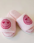 Pink fuzzy slide slippers with a pink heart eyes smiley face graphic on the front of each slipper.