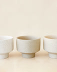 In front of a tan background is three round, ceramic dove pots with a tapered bottom. The pots have black speckles.