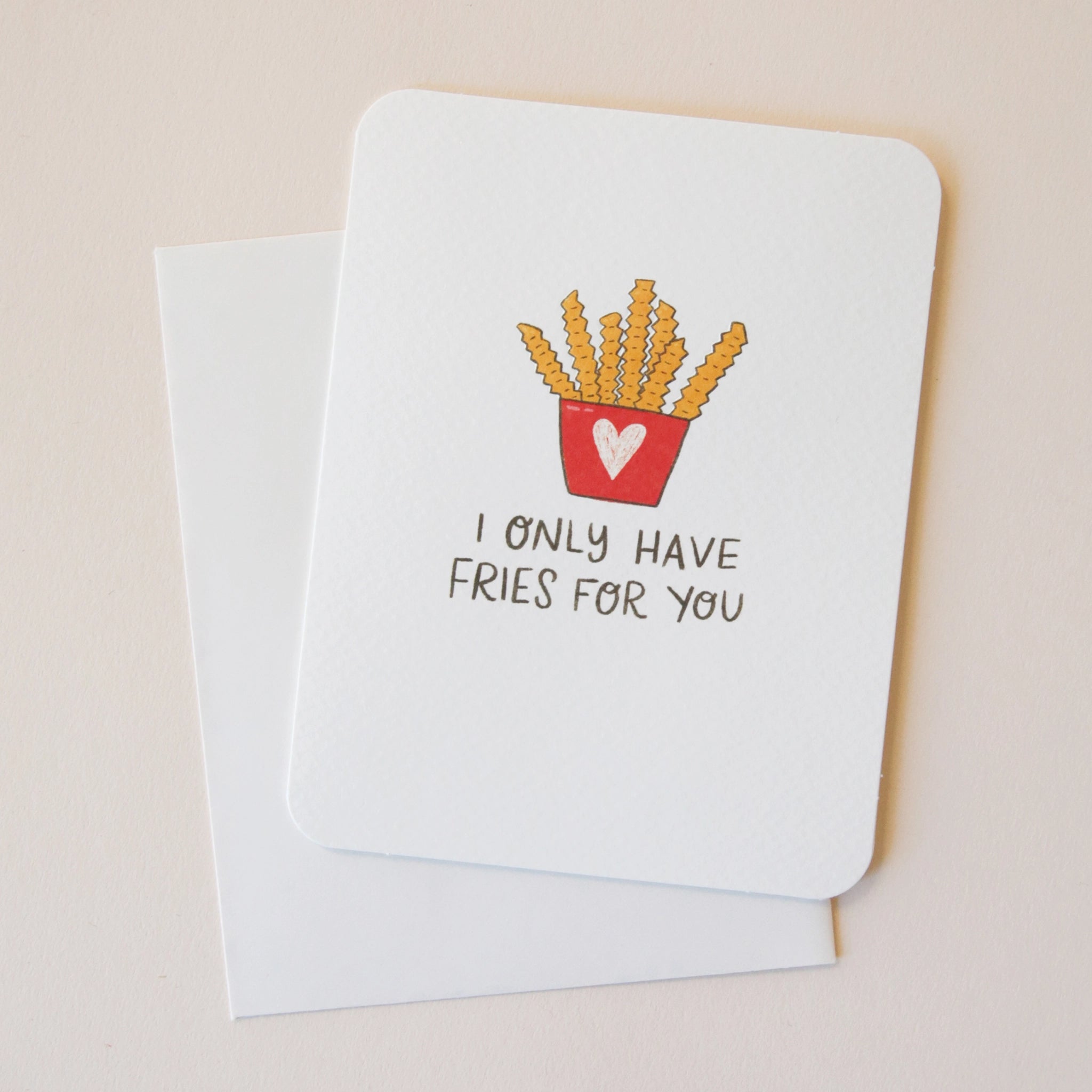 A white card with an illustration of a red carton with a pink heart on the front holding crinkle cut French fries and black text that reads, &quot;I Only Have Fries For You&quot; as well as a coordinating sticker that says, &quot;You&#39;re A Snack&quot; along with a border of fries, popcorn, pretzels and cupcakes.
