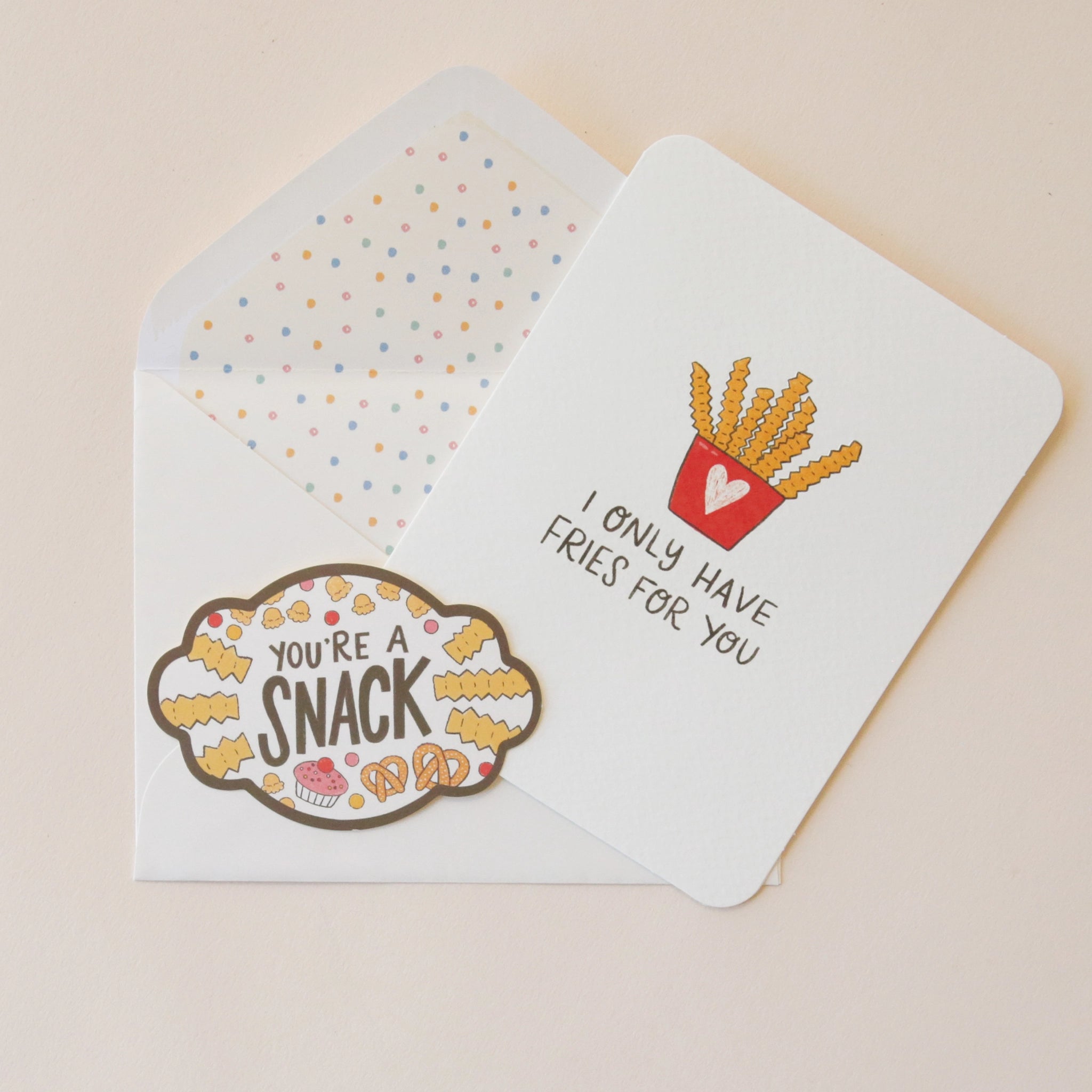 A white card with an illustration of a red carton with a pink heart on the front holding crinkle cut French fries and black text that reads, &quot;I Only Have Fries For You&quot; as well as a coordinating sticker that says, &quot;You&#39;re A Snack&quot; along with a border of fries, popcorn, pretzels and cupcakes.