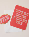 A red card with pink western style text that reads, "You're Doing F*cking Fab" along with a white envelope and a coordinating red sticker that has the same style font and says, "FAB".