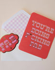 A red card with pink western style text that reads, "You're Doing F*cking Fab" along with a white envelope and a coordinating red sticker that has the same style font and says, "FAB". The interior of the white envelope has a light pink and white checker design.