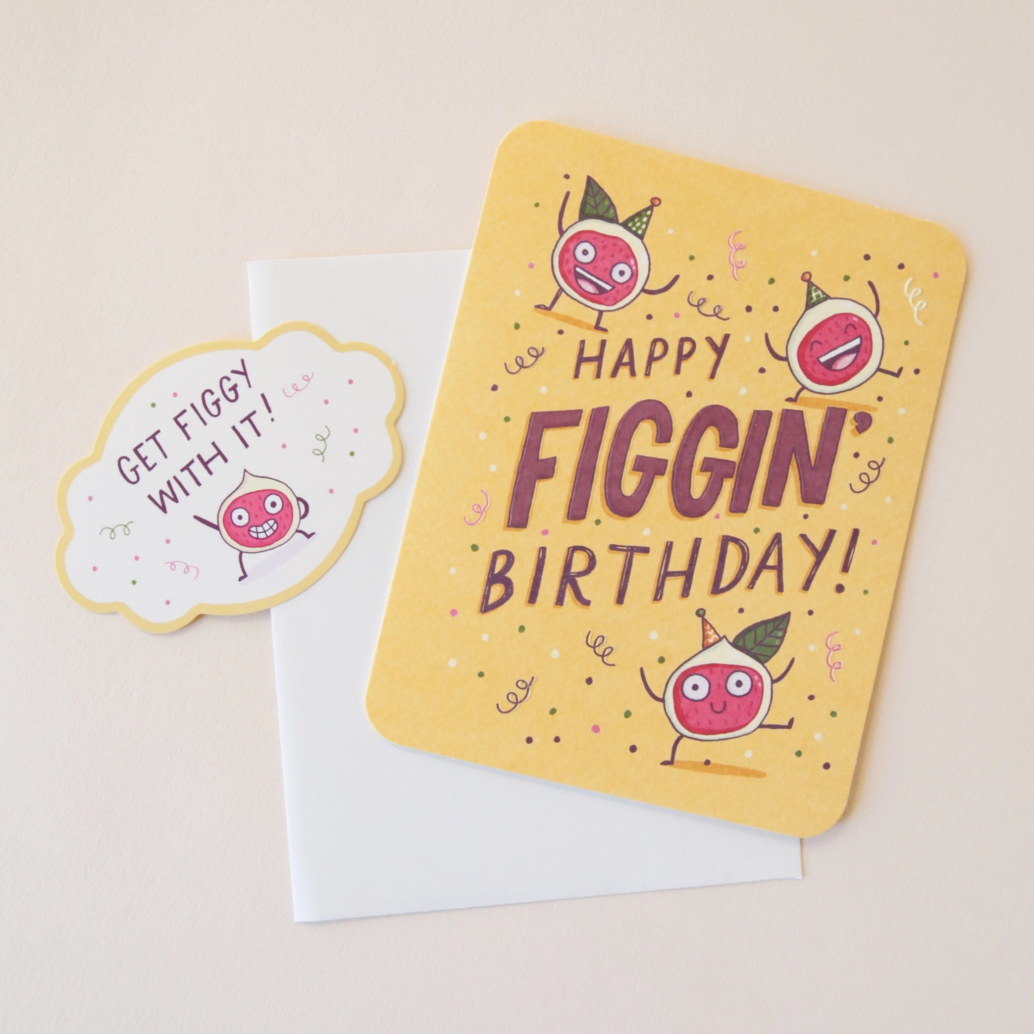 A yellow card with three fig illustrations with their hands up and celebrating along with words in the center that read, &quot;Happy Figgin&#39; Birthday&quot; along with a coordinating sticker that reads, &quot;Get Figgy With It&quot; and a white envelope.