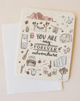 On a neutral background is an ivory card with text in the center that reads, "You Are My Forever Adventure" along with illustrations of various desert camping and road-tripping items, like a camper, a hammock, stars, a campfire and chairs etc as well as a sticker that is included with purchase.