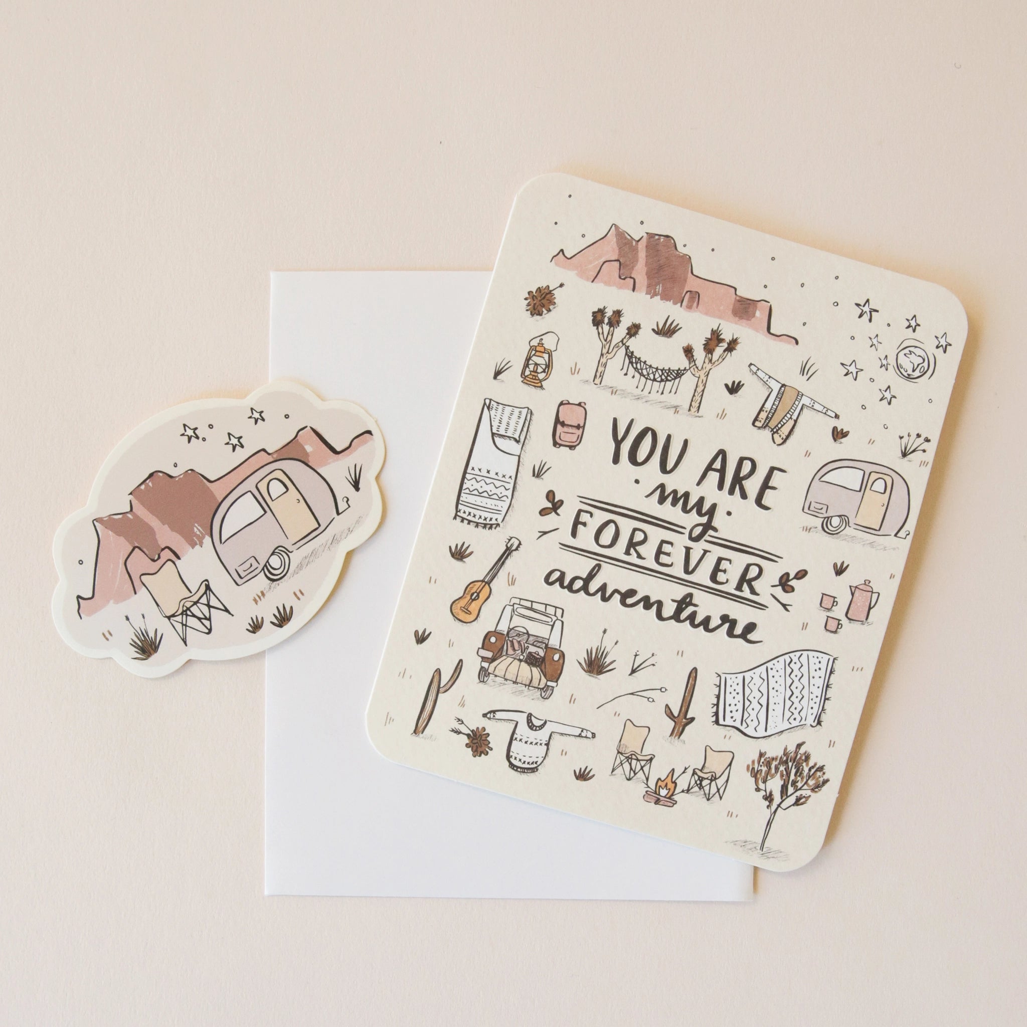 On a neutral background is an ivory card with text in the center that reads, "You Are My Forever Adventure" along with illustrations of various desert camping and road-tripping items, like a camper, a hammock, stars, a campfire and chairs etc as well as a sticker that is included with purchase. 