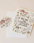 On a neutral background is an ivory card with text in the center that reads, "You Are My Forever Adventure" along with illustrations of various desert camping and road-tripping items, like a camper, a hammock, stars, a campfire and chairs etc as well as a sticker that is included with purchase.