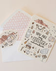 On a neutral background is an ivory card with text in the center that reads, "You Are My Forever Adventure" along with illustrations of various desert camping and road-tripping items, like a camper, a hammock, stars, a campfire and chairs etc as well as a sticker that is included with purchase.