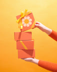 In front of a yellow background is a stack of four orange gift boxes with one VW bus daisy and a yellow bow not included with purchase.