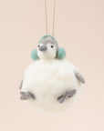 In front of a pink background is a felt penguin ornament. The body is a white, round body. It has two little gray feet and two gray hands. On top is a little white and gray head with a gray nose, two black eyes and green ear muffs.