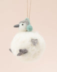 In front of a pink background is a felt penguin ornament. The body is a white, round body. It has two little gray feet and two gray hands. On top is a little white and gray head with a gray nose, two black eyes and green ear muffs.