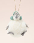 In front of a pink background is a felt penguin ornament. The body is a white, round body. It has two little gray feet and two gray hands. On top is a little white and gray head with a gray nose, two black eyes and green ear muffs.
