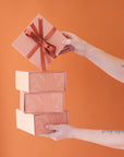 A stack of four salmon pink gift boxes with a magnetic closure and darker pink bird of paradise line graphic along with a rust colored ribbon tied in a bow, not included with purchase.