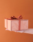 A salmon pink gift box with a magnetic closure and darker pink bird of paradise line graphic along with a rust colored ribbon tied in a bow, not included with purchase.