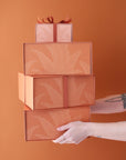 A stack of four salmon pink gift boxes with a magnetic closure and darker pink bird of paradise line graphic along with a rust colored ribbon tied in a bow, not included with purchase.