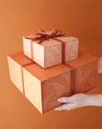 A stack of salmon pink gift boxes with a magnetic closure and darker pink bird of paradise line graphic along with a rust colored ribbon tied in a bow, not included with purchase.