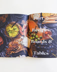 Against a white background is an open book. A picture of the top of a dark table spreads across both pages. In the middle of the picture is a wood tray with cheese, olives, nuts, figs and honey on it. There are plates of crackers on the top right side and plates of salami on the left side. There is white text on the right page that reads ‘part 2: recipes boards & tables.’