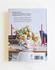 Against a white background is the back cover of a book. The back cover is a picture of a table with a large flower bouquet in the middle. There are red taper candles on both sides of the bouquet. The table is set with dinner plates, silverware and cups. On the top left corner of the picture is black text.