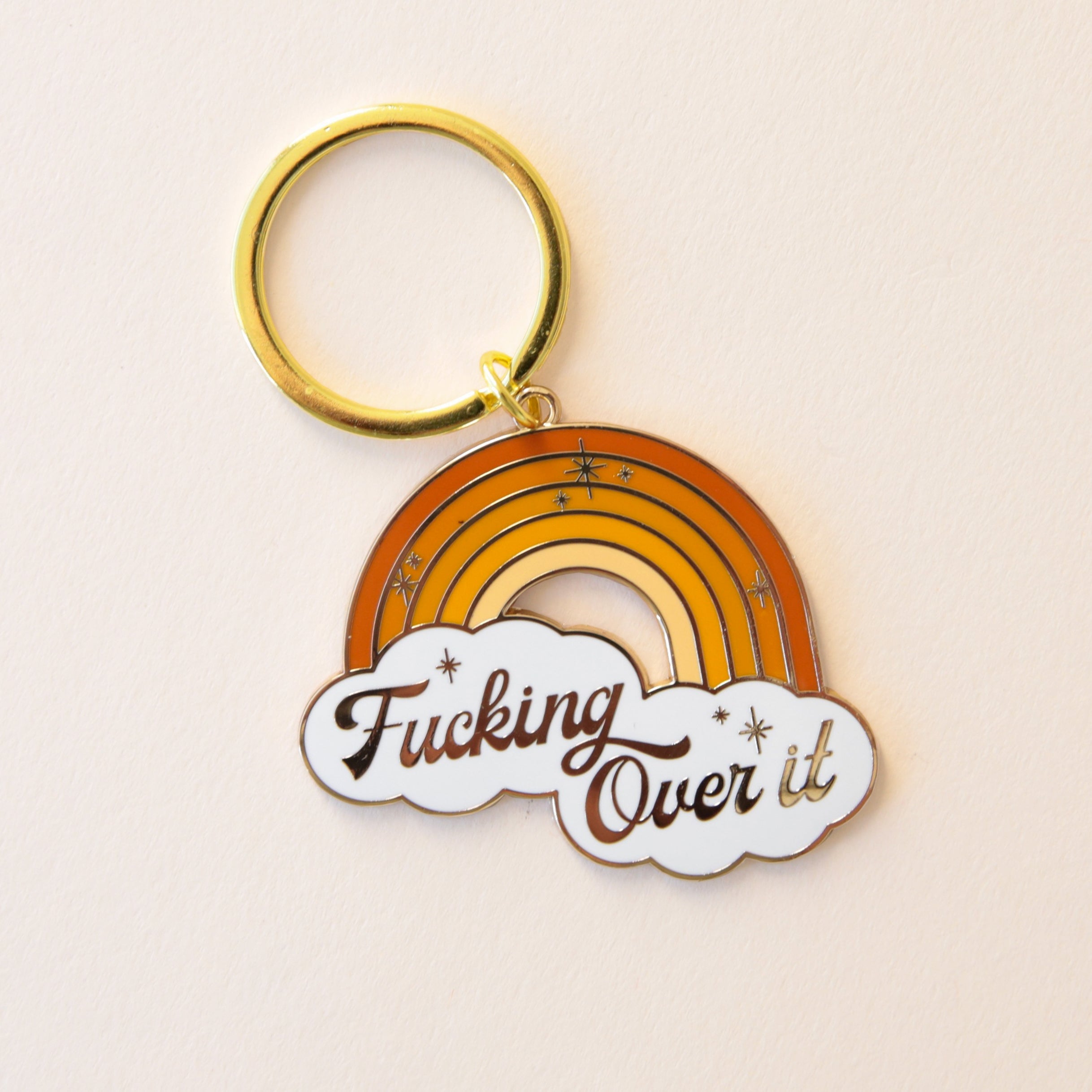A metal rainbow keychain made up of various orange and yellow shades along with white clouds at the bottom and text inside of them that read, &quot;Fucking Over It&quot;. 